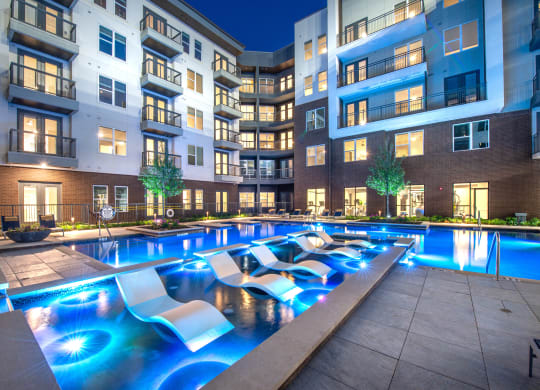 Courtyard pool and tanning area during night time with bright lights in water and view of surrounding apartments and balconies
