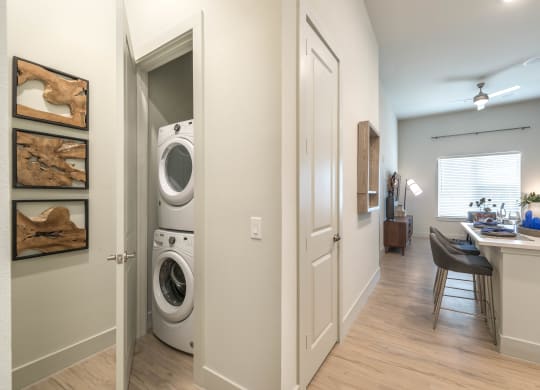 Small room with stacked white washer and dryer and kitchen and living area to right of photo
