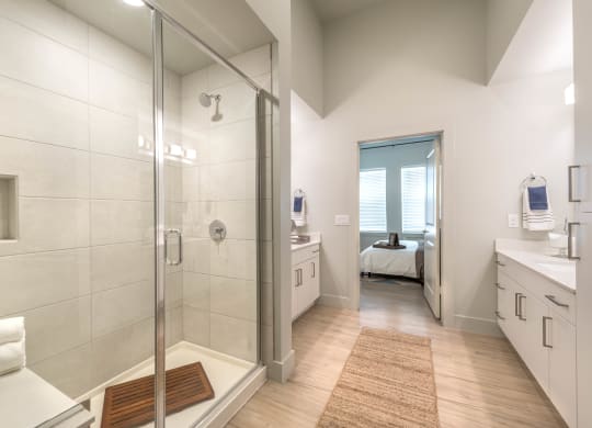 Tiled shower with glass door and a bench with two sinks with white cabinets and countertops on each side of photo with direct view of entrance to a bedroom