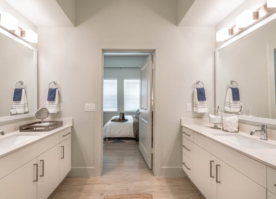 Two sinks with white cabinets and countertops on each side of photo with direct view of entrance to a bedroom