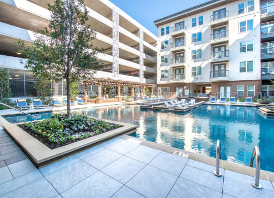 Pool courtyard with pool in the middle and chairs around pool with parking garage behind the pool and a tree surrounded by flowerbed with view of apartment building and balconies