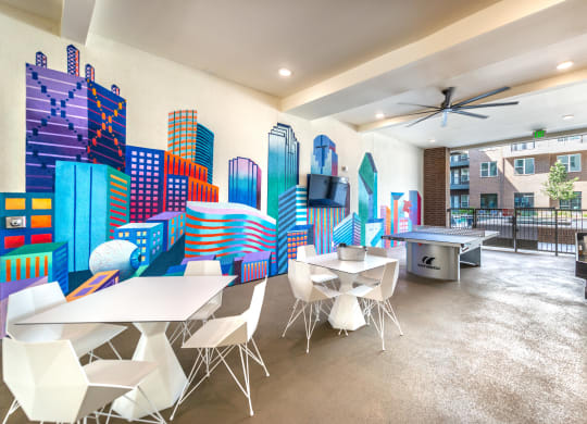 Outdoor covered patio with vibrant painting of Dallas skyline buildings covering wall with two sets of white tables and chairs with ping pong table and shuffleboard on the right side