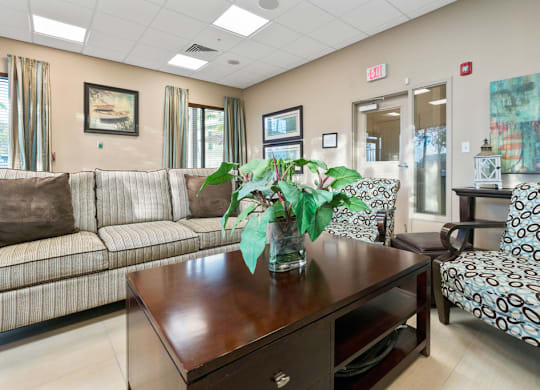 A room with a brown sofa, a patterned chair, and a wooden table with a vase of flowers on it.
