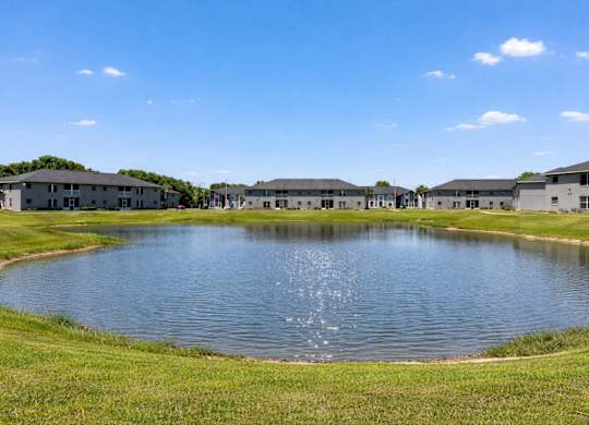 A large pond in the middle of a grassy field with buildings in the background.