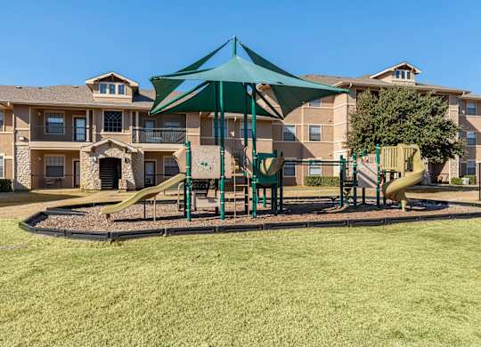 A playground with a green canopy and a yellow slide is in front of apartment buildings.