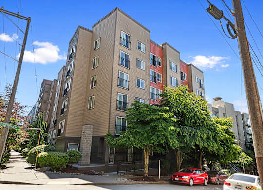 Kitty Corner View of apartment building on a street corner with cars parked, blue skies, and large green trees at Illumina Apartment Homes, Seattle