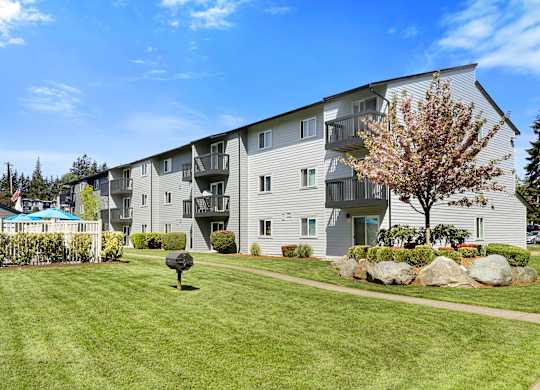 a grassy area with a tree in front of an apartment building at Pacific Park Apartment Homes, Edmonds, WA