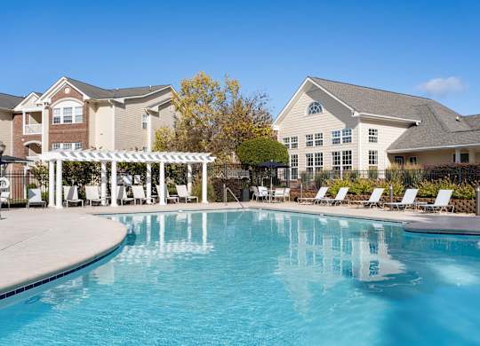 A large swimming pool in front of a building with a white pergola and lounge chairs. at Village on the Lake Apartments, Apartments, Spring Lake, NC
