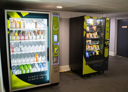 Two vending machines, one with a green and yellow color scheme, the other with a black and yellow color scheme, are in a room with a grey floor and a white ceiling.