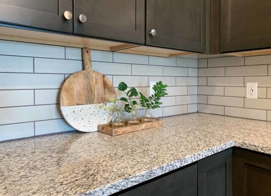 a corner of the kitchen with dark cabinets, a white tile backsplash, and a decorative cutting board