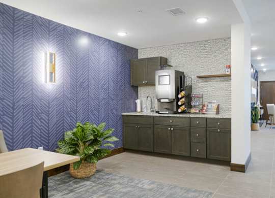Resident coffee bar with dark cabinetry, countertop coffee machine, and open shelving, accented by patterned wallpaper, tile backsplash, and adjacent lounge seating area.