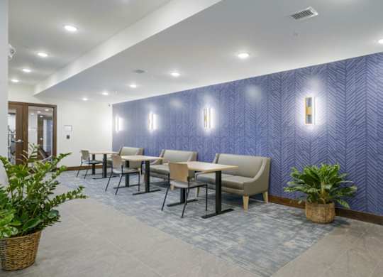Cozy resident lounge seating area with small tables, upholstered chairs and bench seating, patterned accent wall, wall sconces, and indoor plants in a modern apartment community.