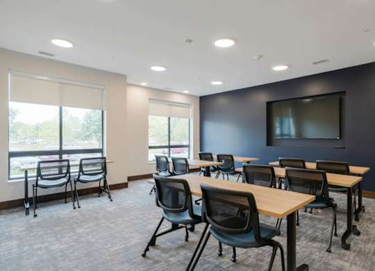 Conference room with light wood tables, mesh chairs, large windows with shades, and wall-mounted TV, offering natural light and open layout.