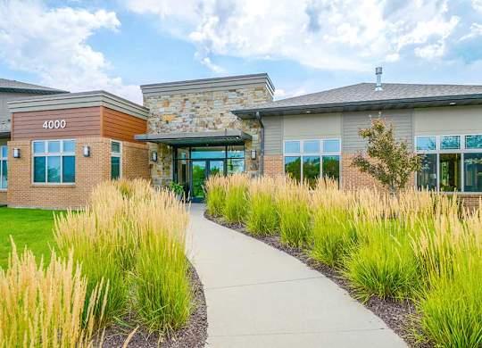 A sidewalk surrounded by tall grass leasing to a modern stone and brick clubhouse with large windows.