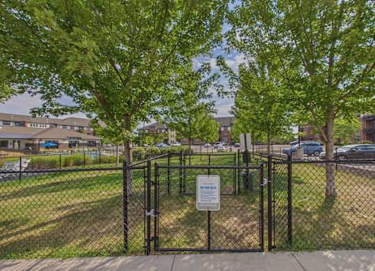 Fenced apartment community dog park with mature shade trees, grassy space, and a gated entrance.