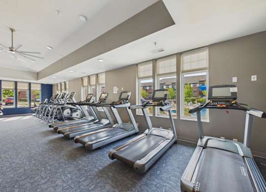 Apartment community fitness center with rows of treadmills and cardio equipment, large windows, and bright overhead lighting.