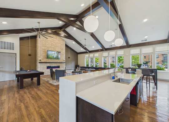 Bright apartment community clubhouse with a large kitchen island, bar seating, pool table, vaulted wood-beam ceilings, and large windows.