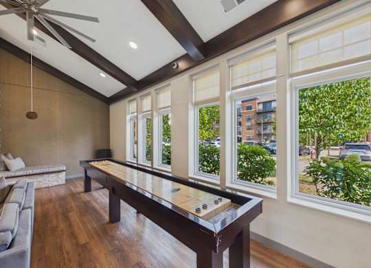 Apartment community game room with shuffleboard table, vaulted wood-beam ceiling, wood flooring, and large windows.