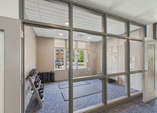 Apartment community fitness room with cardio equipment, rubber flooring, and glass walls overlooking the surrounding property.