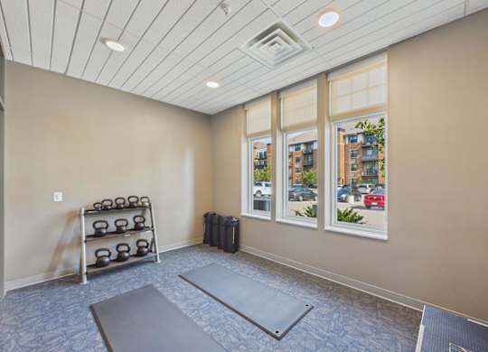 Apartment community fitness area with exercise mats, dumbbell rack, neutral walls, and large windows providing natural light.
