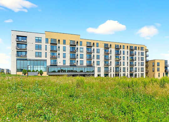 Modern apartment building exterior with multiple balconies, large windows, and mixed-tone facade overlooking open green space.