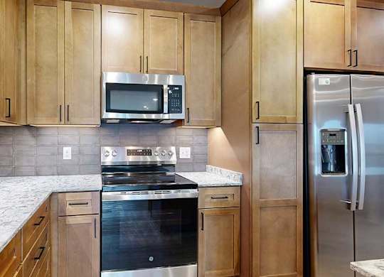 a kitchen with light brown wooden cabinets, quartz countertops, a grey tile backsplash, and stainless steel appliances
