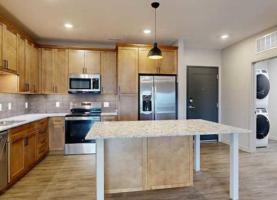 a kitchen with a large table island, light brown wooden cabinets, quartz countertops, and stainless steel appliances
