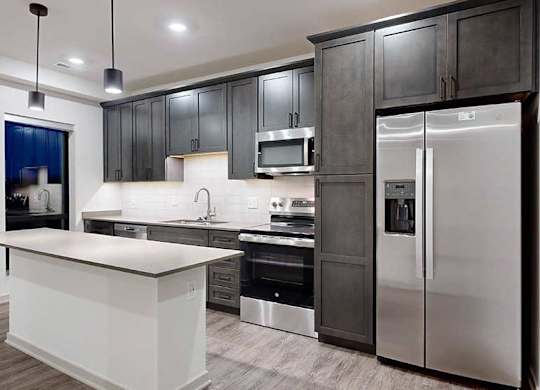 a modern kitchen with dark wood cabinets, light quartz countertops, a narrow central island, a tile backsplash, and stainless steel appliances