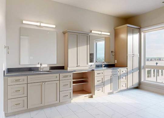 A spacious ensuite bathroom featuring a quartz double vanity with taupe cabinetry, a tall storage cabinet, and tiled flooring.