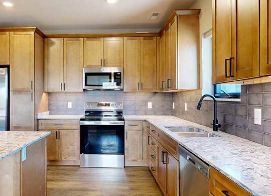 a kitchen with light brown wooden cabinets, stainless steel appliances, a tile backsplash, a large central island, and quartz countertops