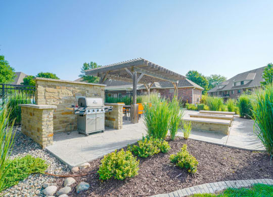 a patio surrounded by greenery with a grill and a fire pit next to a pergola