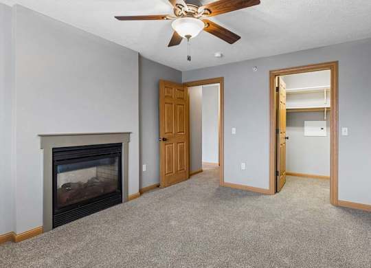 Apartment living room with neutral carpet, a corner fireplace, ceiling fan, and an open doorway leading to a laundry area with shelving.
