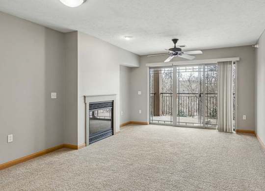 Spacious apartment living room with neutral carpet, a corner fireplace, and sliding glass doors that open to a private balcony with wooded views.