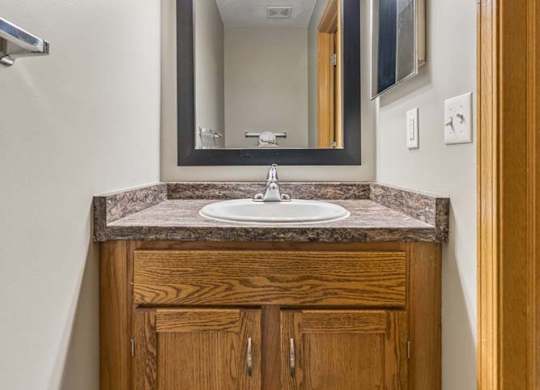 Compact apartment bathroom vanity with a wood-tone cabinet, laminate countertop, framed mirror, modern light fixture, and towel bar in a neutral interior.