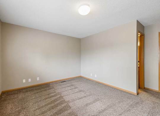 Empty apartment bedroom with neutral carpet, beige walls, wood trim, and an open doorway leading to the adjoining hallway.