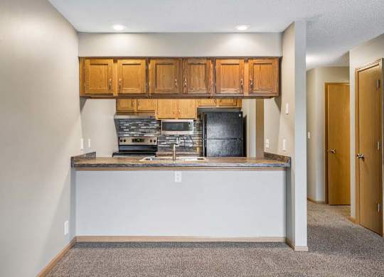 Open apartment kitchen with a breakfast bar, wood-tone cabinets, black appliances, laminate countertops, and a view into the adjacent living area and hallway.