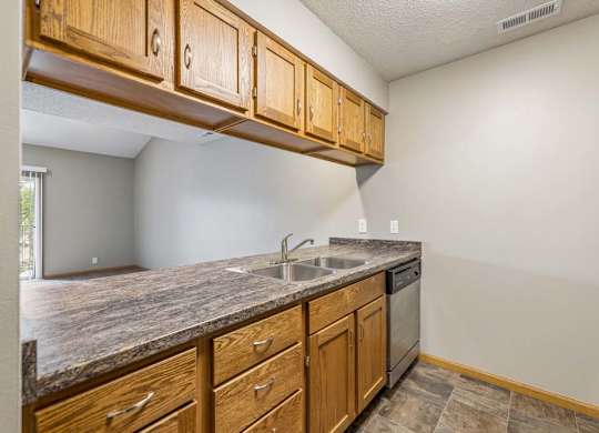 Open apartment kitchen with wood-tone cabinets, laminate countertops, a double-basin sink, dishwasher, overhead storage, and a breakfast bar overlooking the living room.