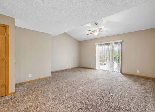 Spacious, empty apartment living room with vaulted ceilings, neutral carpet, a ceiling fan, and a large sliding glass door with vertical blinds that fills the space with natural light.