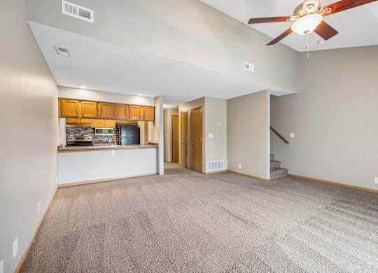 Bright, open-concept apartment living room with neutral carpet, a ceiling fan, breakfast bar overlooking the kitchen, wood-tone cabinets, and a staircase leading to the upper level.