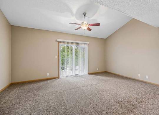 Bright, empty apartment living room with vaulted ceilings, neutral carpet, ceiling fan, and a large sliding glass door with vertical blinds that lets in natural light.