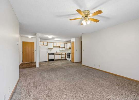 Spacious apartment living room with neutral carpet, a ceiling fan, and an open layout leading to the kitchen with a breakfast bar, wood-tone cabinets, and white appliances.