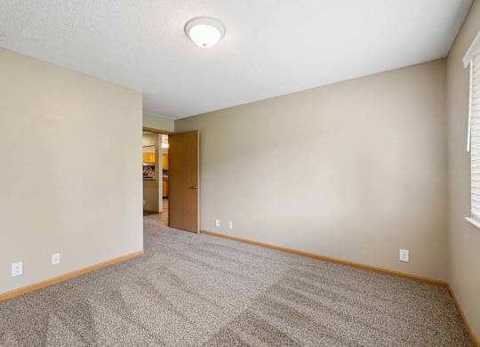 Bright apartment bedroom with carpeted flooring, neutral walls, a window with blinds providing natural light, and an open doorway leading toward the walk-in closet.