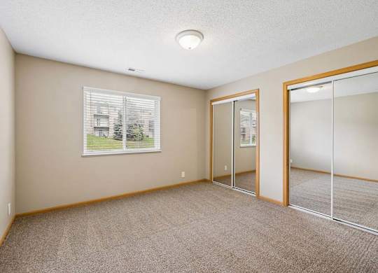 Bright apartment bedroom with carpeted flooring, a window providing natural light, and mirrored sliding closet doors for convenient storage.