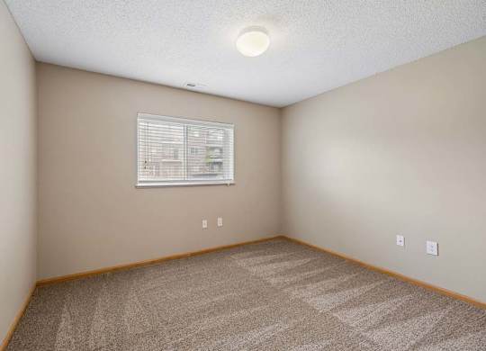 Bright apartment bedroom with carpeted flooring, neutral walls, and a window with blinds providing natural light.