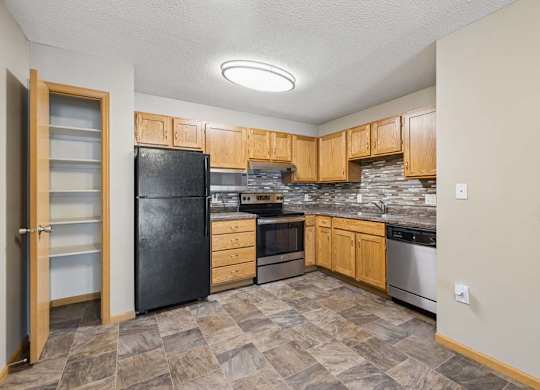 Apartment kitchen with wood cabinetry, a tile backsplash, laminate countertops, black and stainless steel appliances, and a pantry with built-in shelving in a functional U-shaped layout.