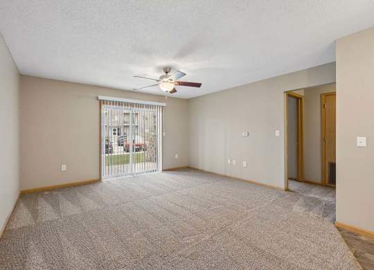 Spacious apartment living room with carpeted flooring, a ceiling fan, and a large sliding glass door with vertical blinds providing natural light and access to a private patio or balcony.