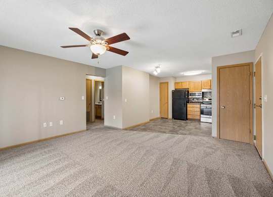Spacious open-concept apartment living room with carpeted flooring, a ceiling fan, and a view into the kitchen featuring wood cabinetry, black appliances, and an adjacent dining area.