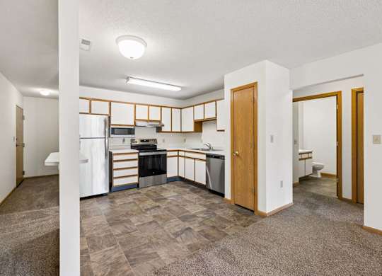 Open-concept apartment with a kitchen featuring white and wood-tone cabinetry, stainless steel and black appliances, a pantry, and adjacent carpeted living and dining areas.