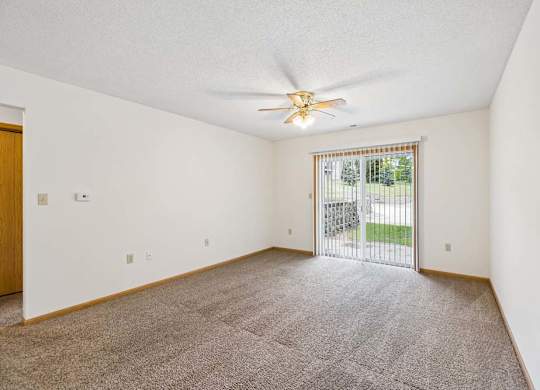 Spacious apartment living room with carpeted flooring, a ceiling fan, and a large sliding glass door with vertical blinds providing natural light and access to a private patio or balcony.