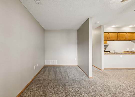 Spacious apartment living area with carpeted flooring and a flexible nook, adjacent to an open kitchen with a breakfast bar and wood cabinetry.